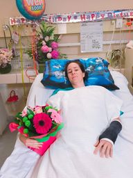 A young woman, Ebonny, lays in a hospital bed on her birthday, following a catastrophic injury accident. She's holding a pink bouquet of flowers  on one arm and a black cast on the other. Birthday banners and balloons are in the background.