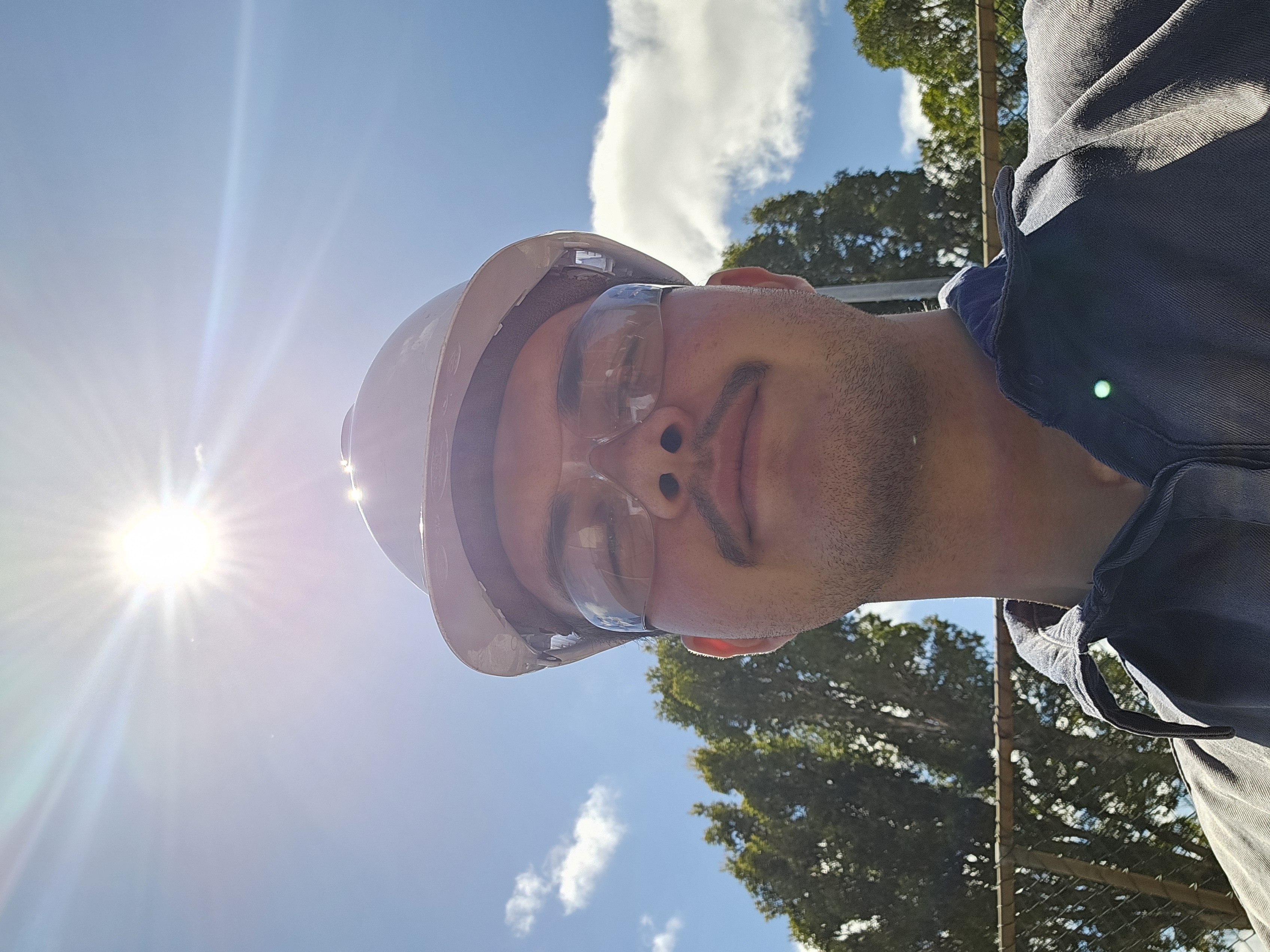 A young man stands with a white hard hat on, facing the camera, wearing safety glasses. The sun shines brightly in the background, against a blue sky with white clouds and green trees.