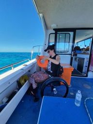 A young woman, Ebonny, sits in a wheelchair on board a boat out at sea. She's holding an orange float and blue skies and water glisten in the background.