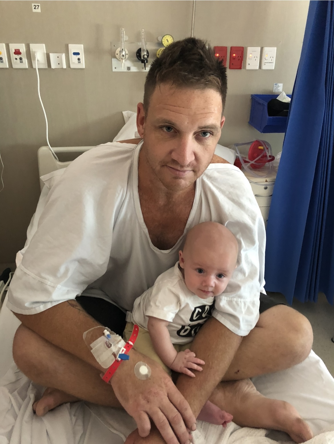 A young man sits on a hospital bed with a full head of hair, in a white hospital gown, holding his young baby.