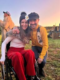 A young woman, Ebonny, sits in a wheelchair dressed in red trousers and a white top. She hugs a man wearing a grey hat with a flower attached, wearing a yellow top and grey trousers. The sky is pink in the background as the sun sets.