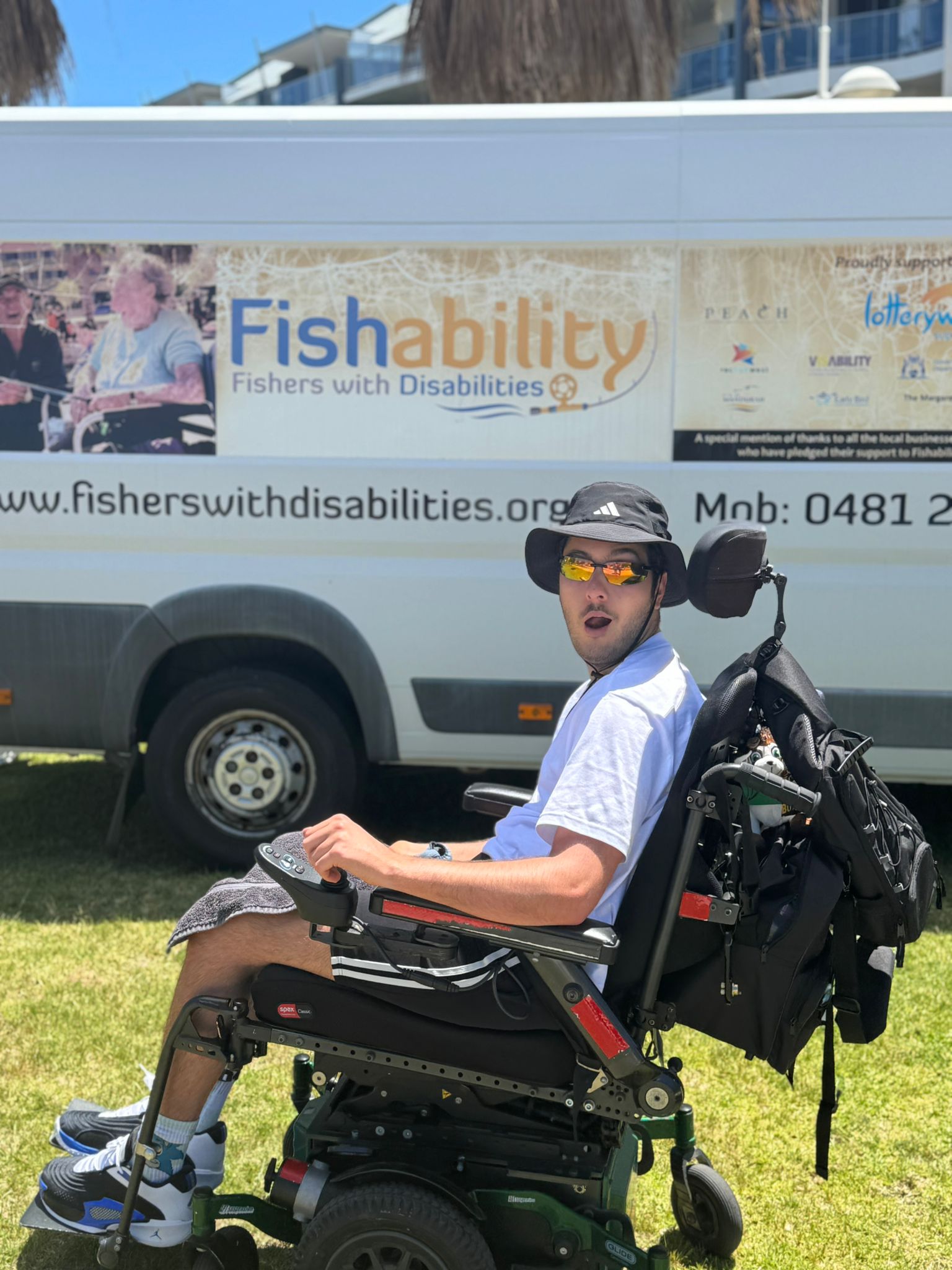A young man, Liam, sits in a black powered wheelchair wearing orange mirrored sunglasses, a black bucket hat, white t-shirt, black shorts, in front of a 'fishability' white van on a sunny day.