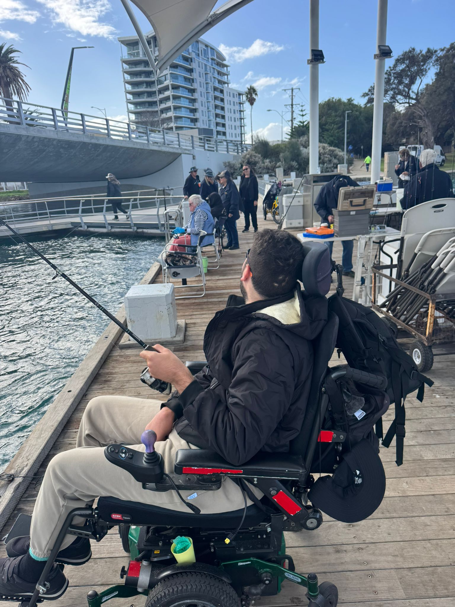 A young man, Liam, sits in a black powered wheelchair on a fishing dock, holding a fishing rod looking away from the camera. He has dark hair. The sky is blue, speckled with some white clouds and the ocean water is teal.