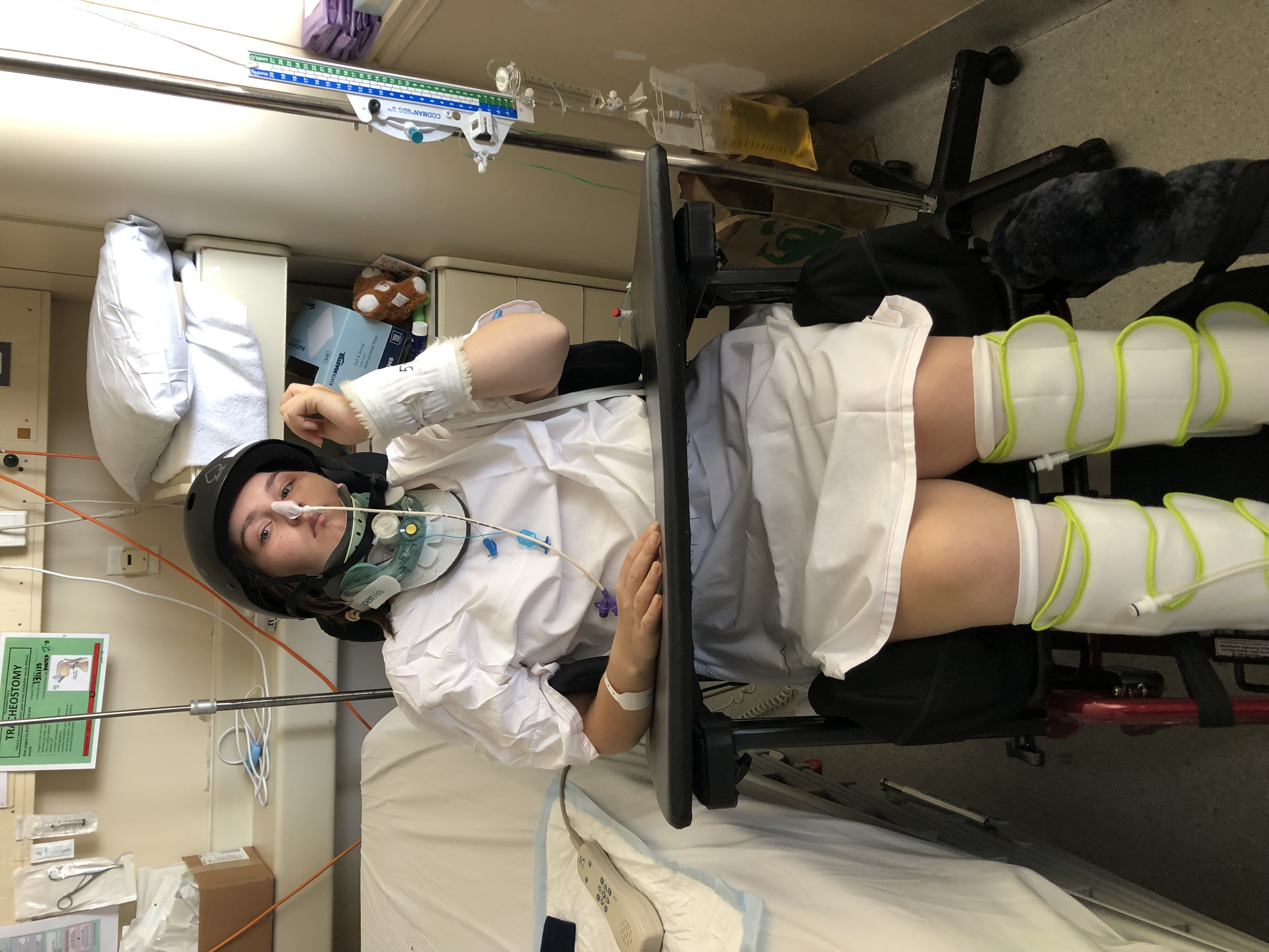 A young woman, Laura, sits in a powered wheel chair beside a hospital bed wearing a crash helmet, neck brace, white hospital gown, and leg braces following a catastrophic injury.