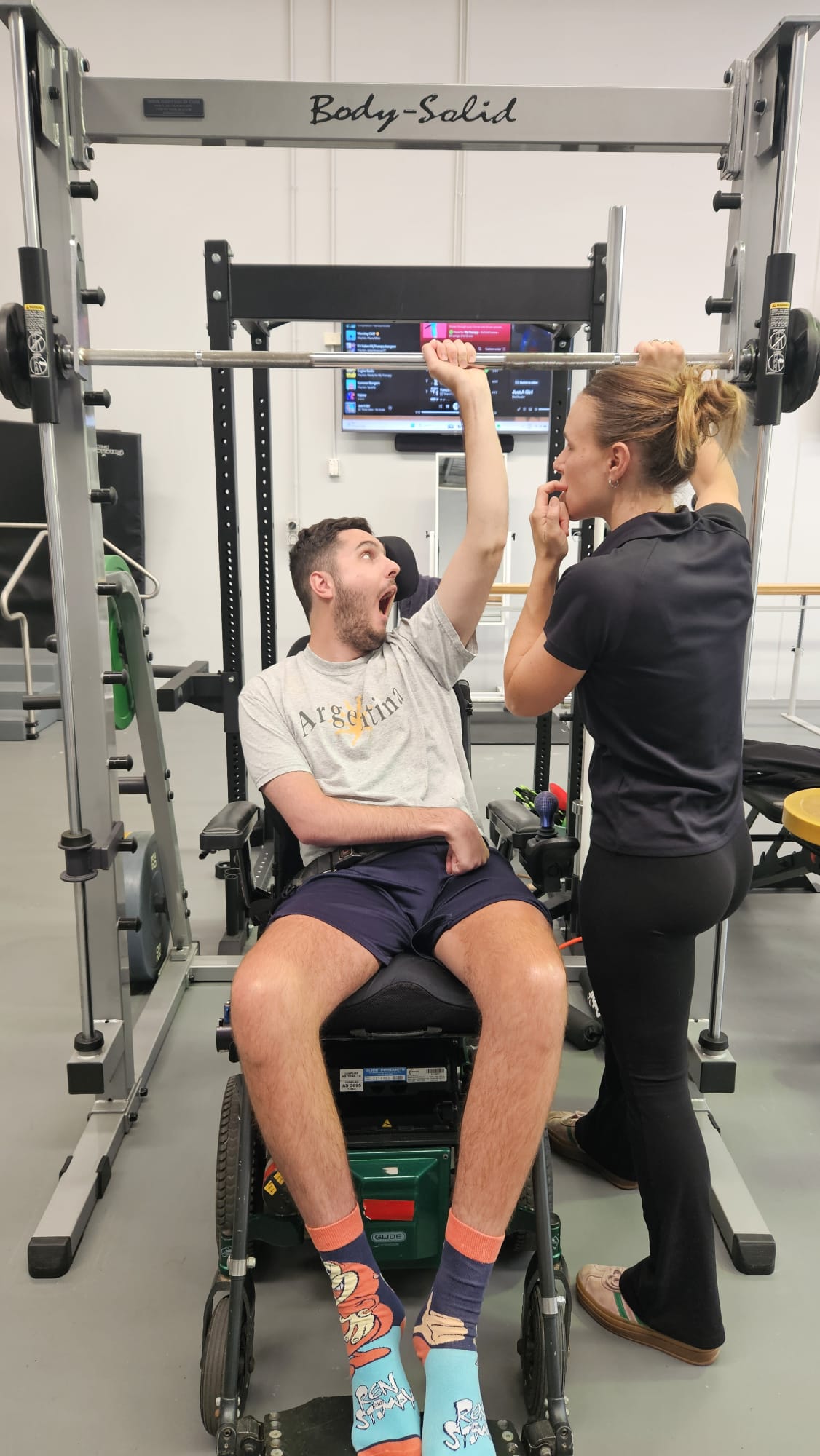 A young man, Liam, sits in a black powered wheelchair wearing a grey t-shirt, navy blue shorts. He's in a gym, with a female physiotherapist by his side. Is is lifting weight with one of his arms, with the support of the physiotherapist.