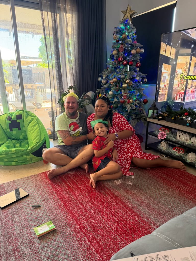 A man, Dylan, sits with his wife, Ana and young son, Lenny, in front a Christmas tree on a red rug.