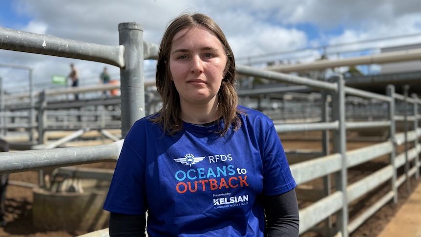A young woman, Laura, wears a blue 'Oceans to Outback' tshirt stood in front of a gated cattle pen. The sky is blue and full of white clouds in the background.