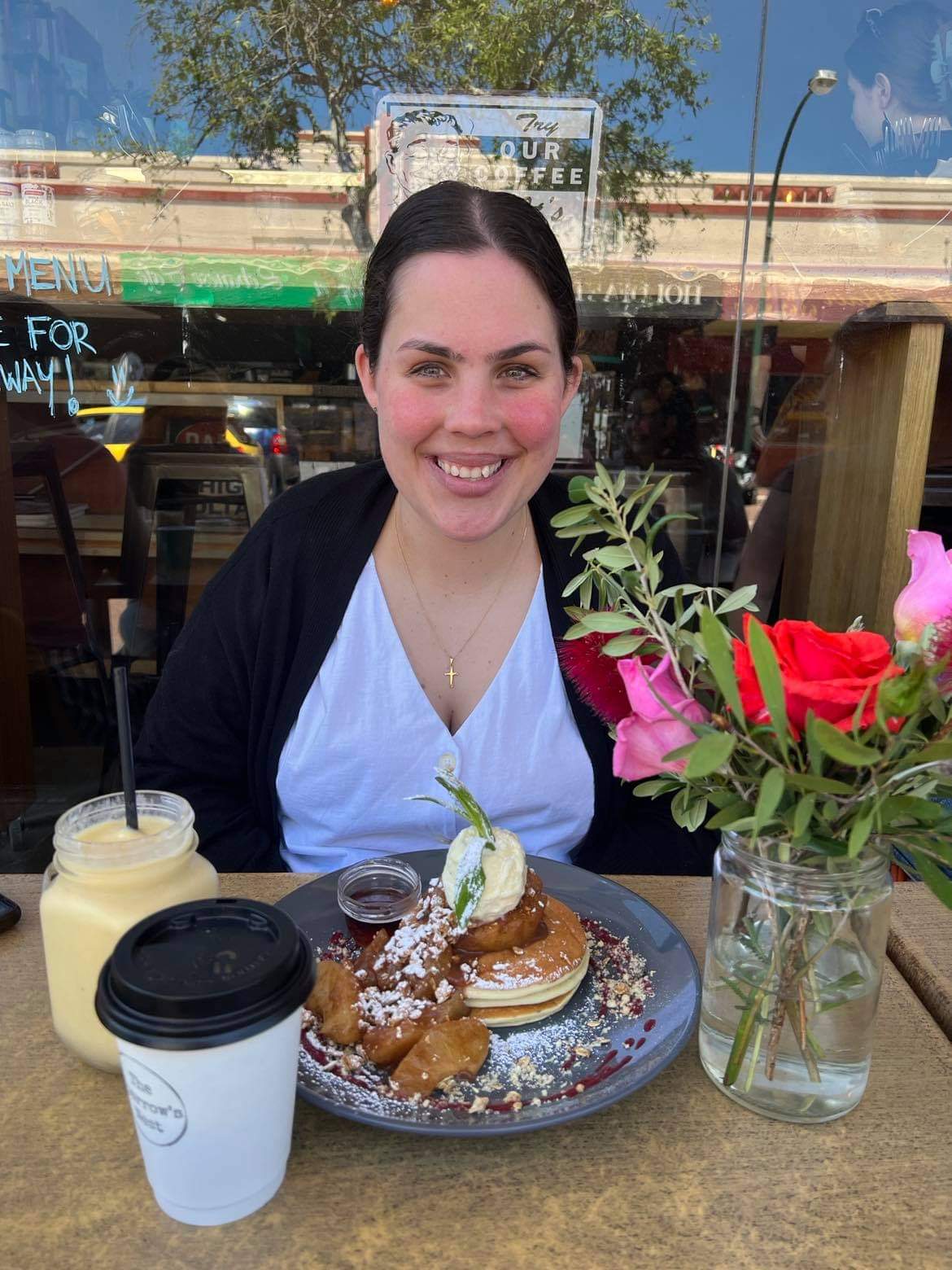 A young woman, Liz, sits at a restaurant table outside smiling facing the camera, with a smoothie, coffee and plate of pancakes. There's a vase full of red and pink roses sat on the table in front of her.