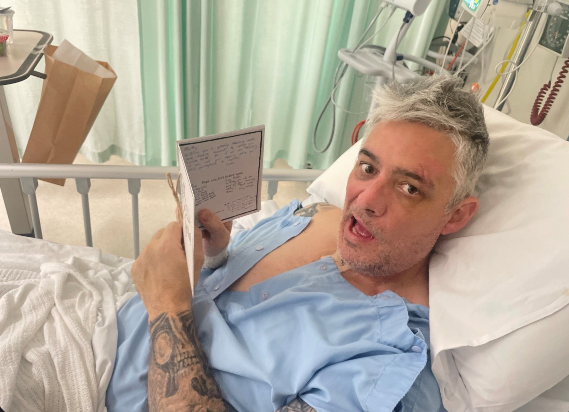 A man, Matt, lays in a hospital bed in a blue hospital gown. He's holding a card and looks directly at the camera. He has grey hair and tattoos on his arm.